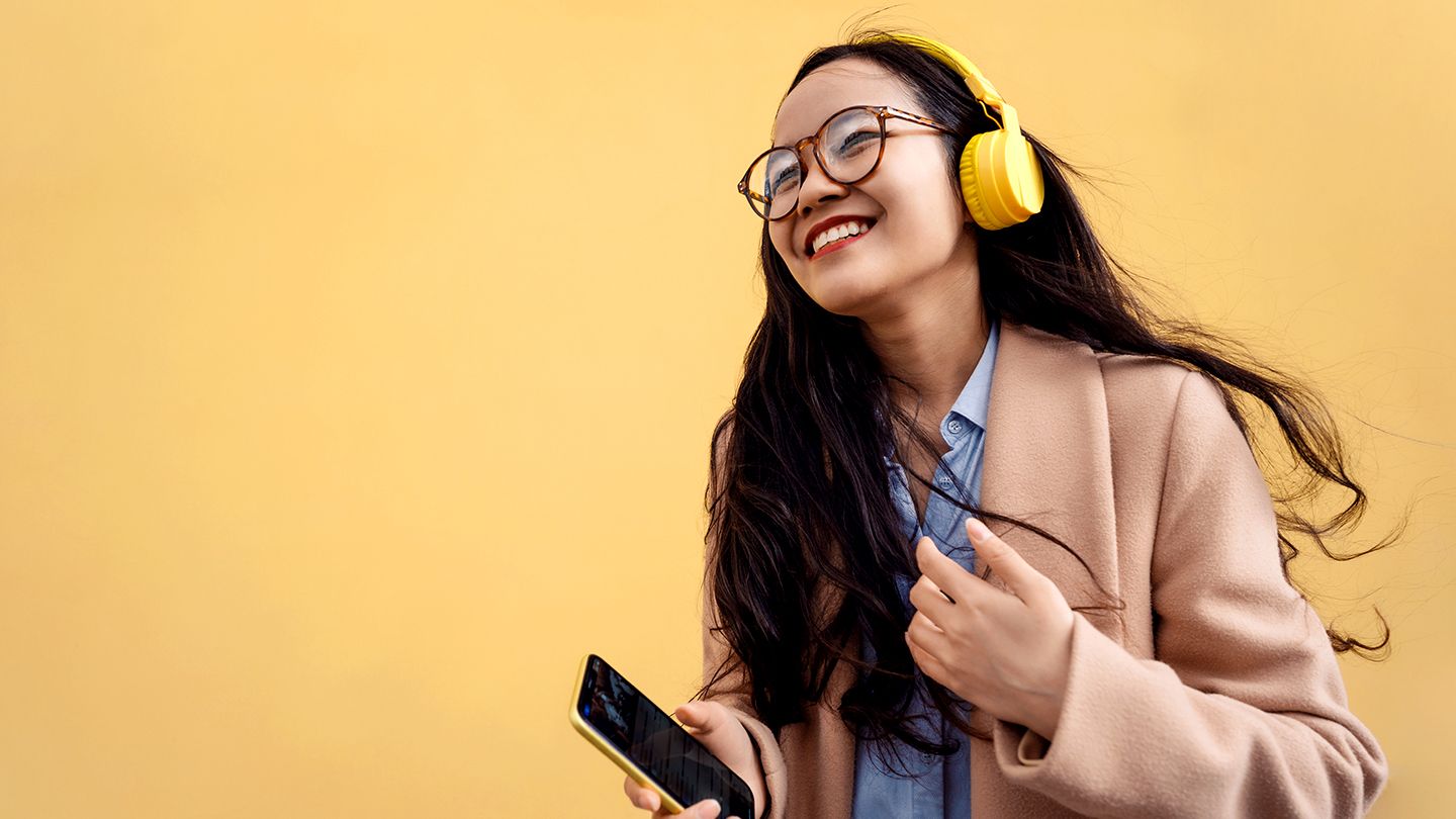 an orange background with a girl wearing spectacles, blue shirt and peach coloured coat, yellow headphones and holding a mobile